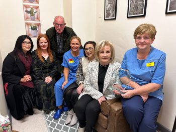 Six females and one male sitting on sofa with one being handed an award