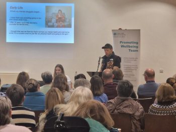 A man speaking to a crowd in front of an audience. A presentation is projected on the wall.