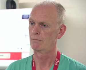 A man in light green scrubs and a red lanyard is talking to someone out of shot.