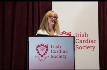 A woman is speaking at a lectern. The lectern and banner behind read "Irish Cardiac Society" and have the logo to the left of the name.