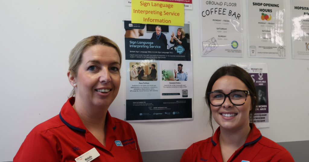 Two Emergency Department staff are pictured beside a poster promoting access to sign language interpreting services. The poster outlines face-to-face and remote interpreting options and is displayed on a hospital noticeboard with other patient information notices.