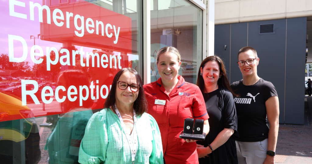 Four people stand outside the Emergency Department reception entrance. One person holds two handheld pager devices beside a large red sign reading “Emergency Department Reception
