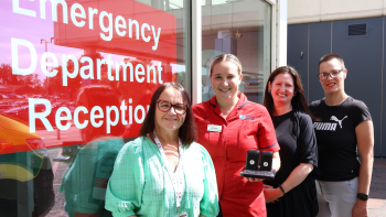 Four people stand outside the Emergency Department reception entrance at a hospital. One person in a red uniform holds two handheld pager devices as the group poses beside a large red sign reading “Emergency Department Reception